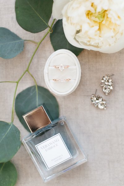 A flatlay of wedding details, including rings, earrings, and perfume with floral details.