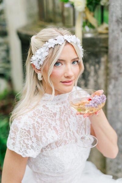 A bride holds a cup of champagne with florals up to her mouth. 