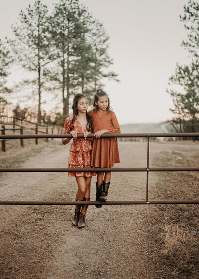 Two sisters wearing cowgirl outfits and their cowgirl boots stand close together on the bottom rung of a cattle gate with pine trees in the background.