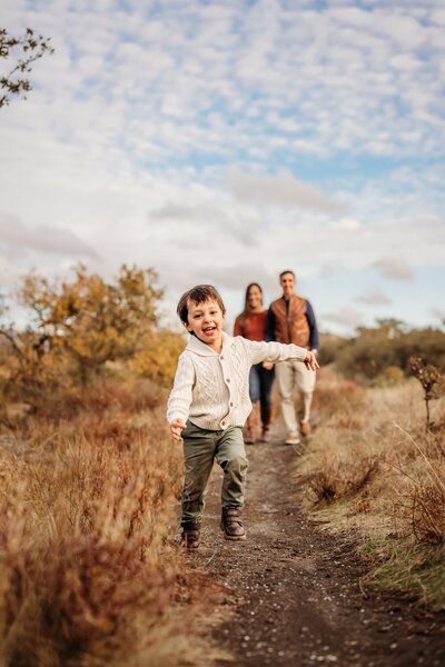 happy toddler runs towards the camera as his parents walk behind him hand in hand