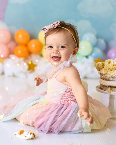 A smiley baby is sitting in front of rainbow balloons, enjoying her cake smash