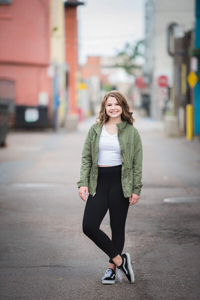 A woman in a green jacket and black pants stands in an alley smiling