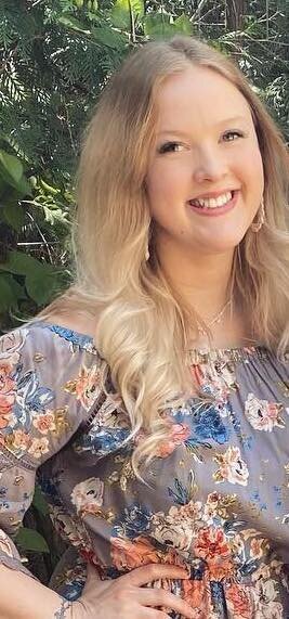 A blonde woman wearing a gray floral shirt is smiling at the camera.