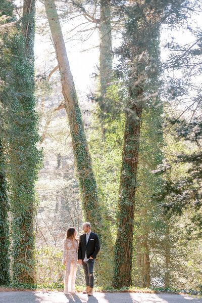 Bride wearing an ivory lace jumpsuit holds hands with her groom wearing boots and black pants and a black jacket and looking at each other in front of giant trees in the Biltmore Forest in North Carolina.