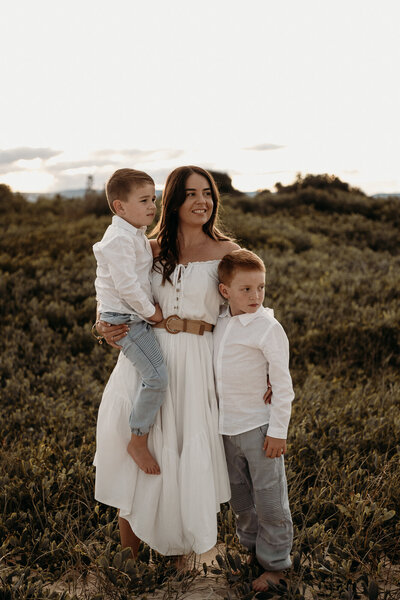 A family of five, including two adults and three children, stands together outdoors during sunset, smiling for the camera. The background features tall grass and a hilly landscape—expertly captured by a South Coast family photographer.