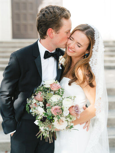 Close up image of groom kissing bride on the cheek while she smiles holding her bouquet