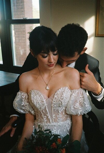 A couple shares an intimate moment indoors during their wedding in Tucson, Arizona. One partner, wearing an off-the-shoulder lace gown, looks down while the other, dressed in a suit, gently embraces them from behind, their foreheads almost touching.