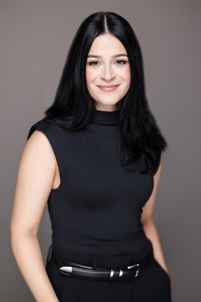 Corporate headshot of a woman with long black hair wearing a black top and belt on a gray background, Long Island professional headshots.