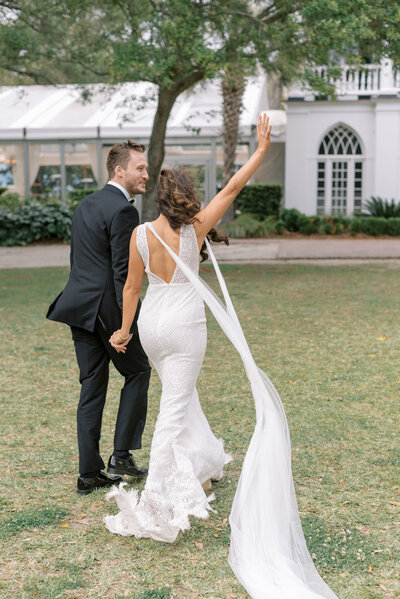 Bride and groom holding hands as the bride's cape blows in the wind in front of the house at Lowndes Grove in Charleston South Carolina