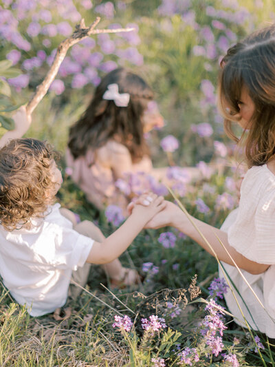 Mother holds toddler son's  hand while they and older sister sit in a field of purple wildflowers taken by Little Rock family photographer Bailey Feeler Photography