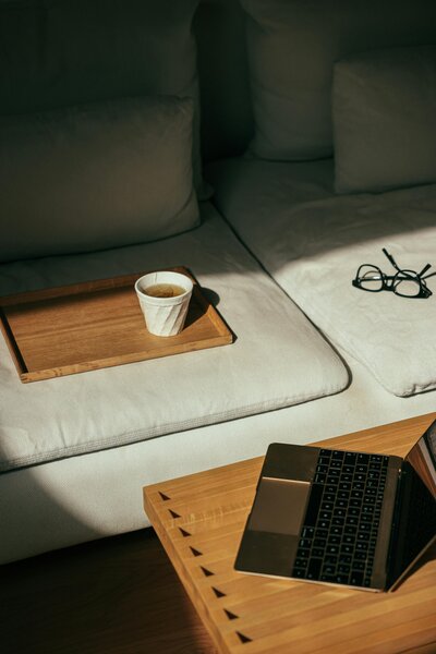 Minimalist home workspace with a laptop on a wooden table, soft neutral seating, glasses, and tea — a calm setup for writing Shopify blog content and bright ideas.