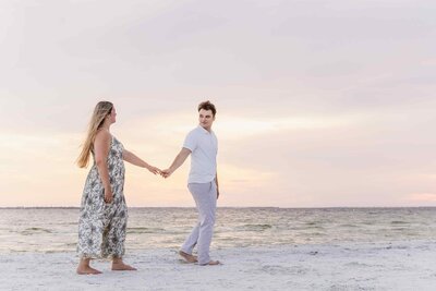 Man leads his new fiancee along the beach with a pastel sunset