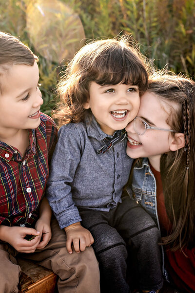 Three young children sitting in a grassy field smiling at each other.