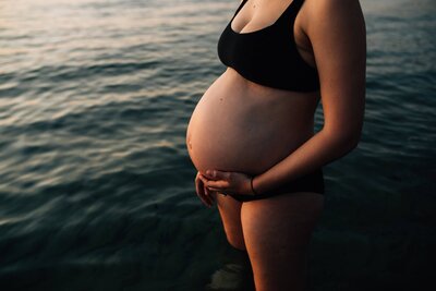 Pregnant woman in black bikini standing in water at beach.
