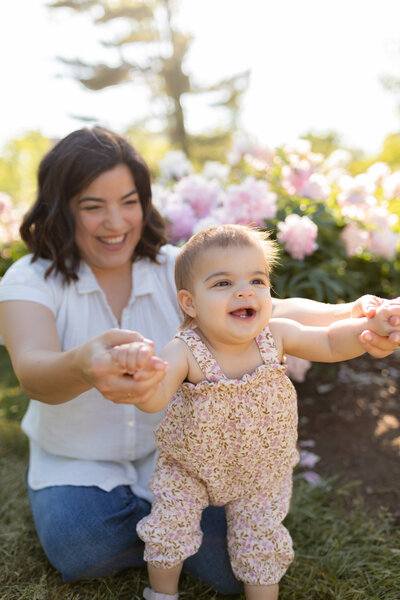 mother leads baby daughter with hands as she learns how to walk at the peony gardens at nichols arboretum in ann arbor michigan