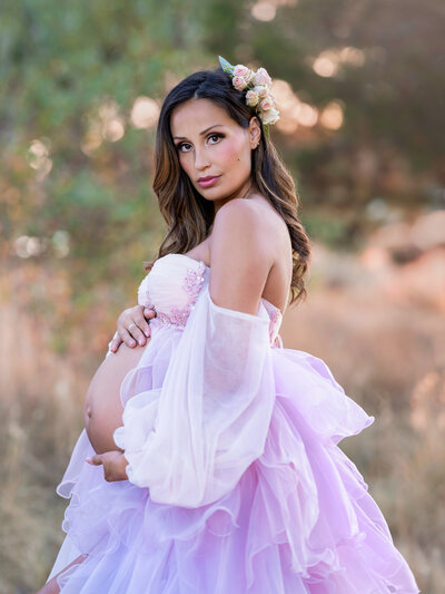 Pregnant woman wearing a pink and purple elegant maternity dress and a floral accessory in her hair standing in front of some foliage while holding her belly with one hand above and one hand below her pregnancy bump.