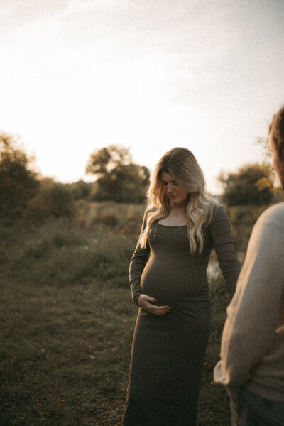 A woman looking down holding her pregnant belly at sunset in a field | Durham, Ontario