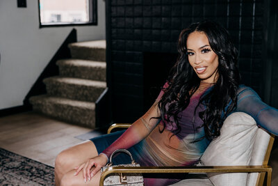 Tiara in a multicolored dress sitting in a white and gold chair in front of a black fireplace