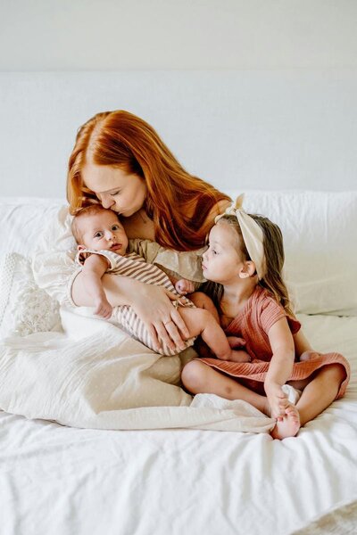 Mother kissing her newborn baby while the older sibling looks on lovingly, capturing a tender family moment.