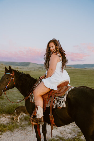 A woman in a white dress sits on a horse in a pasture at sunset