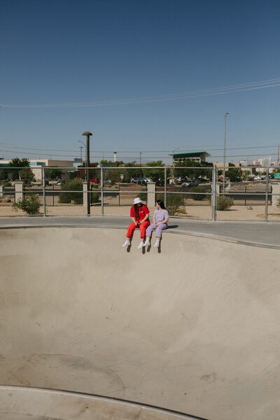 Two girls sitting on the edge of the bowl at the skate park.