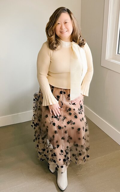 Anne, an Asian woman, sitting on a chair, wearing a light-colored top and a flowy, floral skirt.
