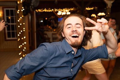 A wedding guest is dancing while making peace signs at the camera during  the wedding reception.