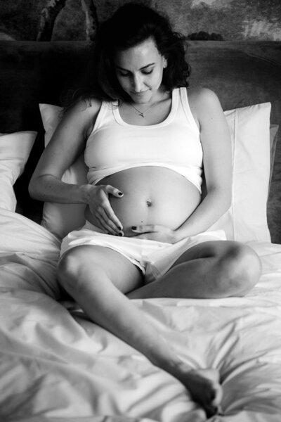 Pregnant woman sitting on a bed in soft light, cradling her belly with both hands.