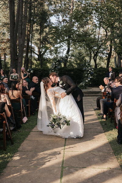 Groom dips the bride into a kiss at the end of the ceremony aisle as guests cheer and clap during an outdoor forest wedding.
