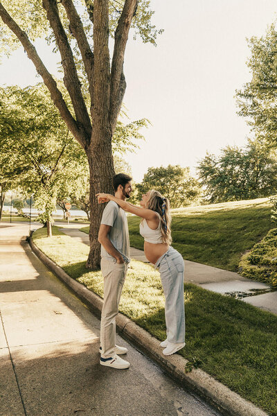 Expecting mother stands on curb with arms draped around her husband's shoulders