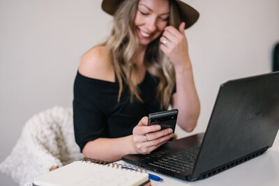 Sammy smiling sitting at her desk with a laptop holding her phone