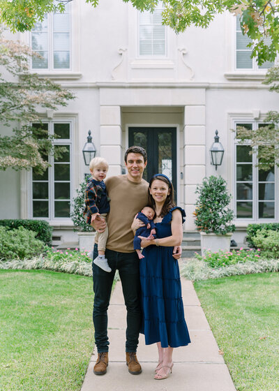 Family with a toddler and newborn boy standing in front of a luxury home in Dallas that is framed by tree leaves.