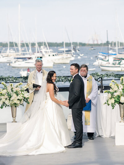 A Jewish-Christian couple holds hands during their wedding ceremony on a waterfront. The bride wears a white gown, and the groom is in a black suit. Two officiants stand nearby. Boats and a scenic body of water are visible in the background.