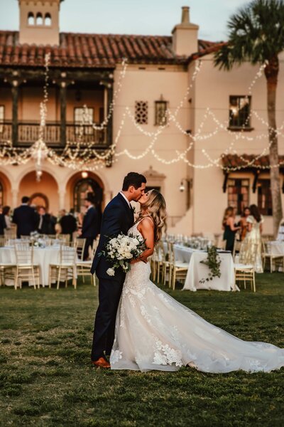 Shannon Lee, an Orlando wedding photographer capturing a romantic moment between a couple sharing a kiss in front of a beautifully lit venue, with their wedding reception in the background.