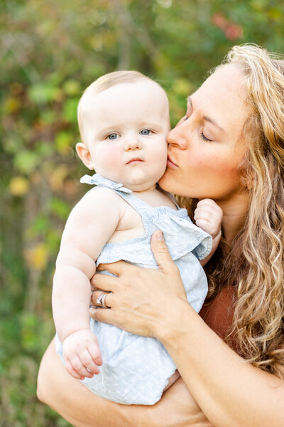 a mother kisses her toddlers cheek while the toddler looks straight at the camera
