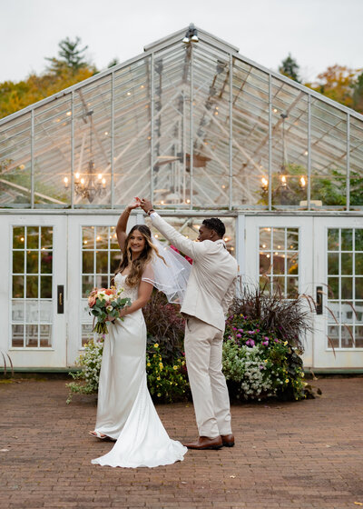A bride and groom dance outside a large glass greenhouse. The groom twirls the bride, who holds a bouquet of flowers. The bride wears a white gown and veil, while the groom is in a light beige suit. Surrounding them are lush green plants.