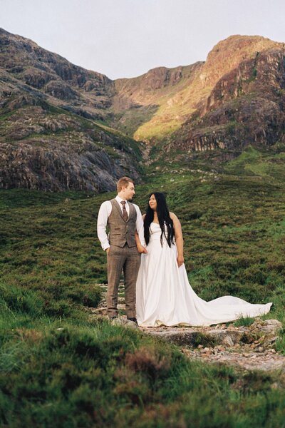 A 35mm film photograph of a couple standing side by side underneath a giant Aberdeenshire mountain. They wear wedding attire. 