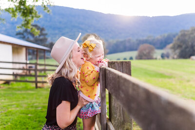 Mother kisses toddler daughter climbing on the wooden fence at Mountain Cove Farms in Chickamauga, Georgia