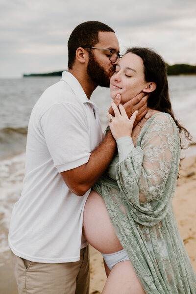 Husband kissing pregnant wife's cheek on the beach in Maryland.