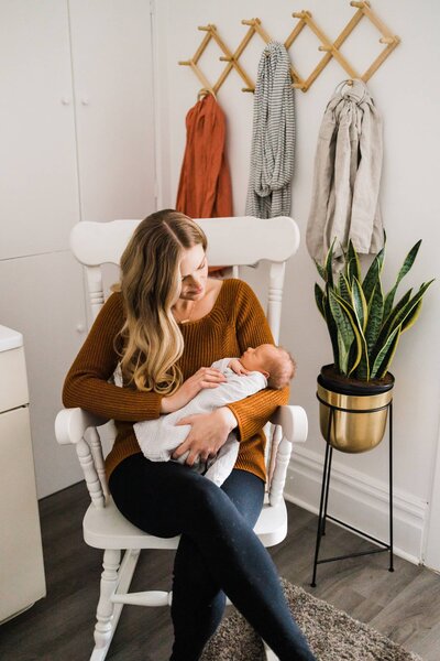 Woman sitting in a white rocking chair, lovingly holding and looking at an infant in a cozy room, representing the essence of newborn lifestyle photography.