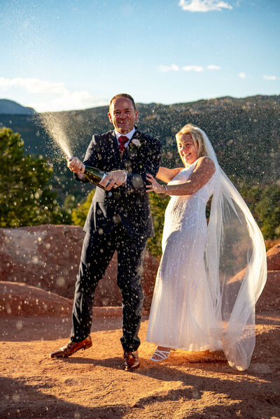 Bride and groom pop a bottle of champagne and spray it towards the camera. The sun reflecting off the champagne spray. The couple is laughing.