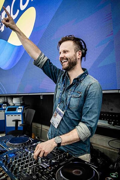 Smiling male DJ in a denim shirt raising a hand mid-performance at a tennis stadium with United Cup branding