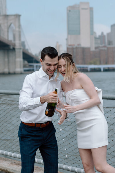A couple celebrates with champagne by a river with a cityscape background, sharing a toast in casual wedding attire.