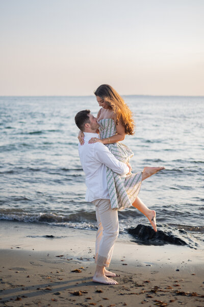 Collins Beach Newport RI | Kelsey Sheehan Photography Timeless Rhode Island Weddings | A man lifts a woman joyfully near the ocean at sunset. Both are smiling, and she is barefoot in a striped dress. The mood is romantic and serene.