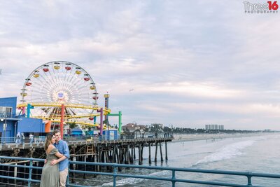Engaged couple embrace while standing on the Santa Monica Pier for engagement photos with the Ferris Wheel in the background