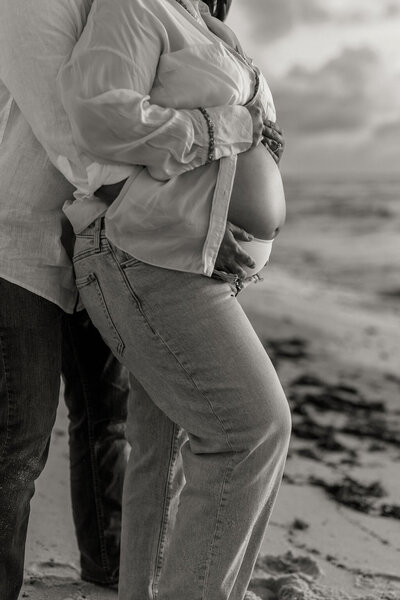 Black and white close-up of pregnant belly with hands gently resting, captured during a maternity session on the beach in Miami.