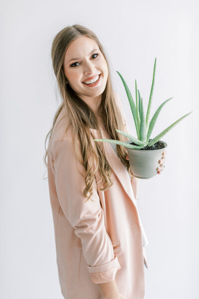 A photographer wears a pink jacket while holding a plant in front of a white wall.
