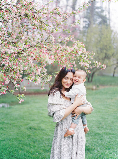 A mother embraces her infant son while standing near cherry blossom tree in Brookside Gardens in Maryland