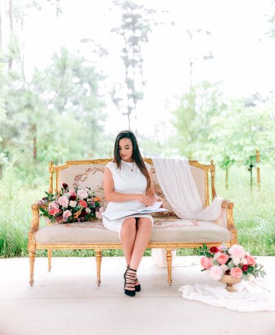 Event planner Linda Linares seated on an ornate gold settee with pink florals, writing in a notebook at an outdoor venue.
