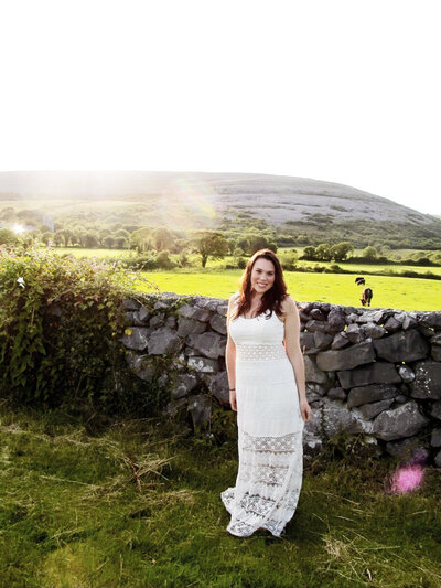 Woman smiling, wearing a white dress standing in front of a stone wall in a filed